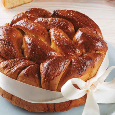 Bolo trançado dourado com laço branco em prato azul, favo de mel de madeira e fatia de bolo na mesa clara