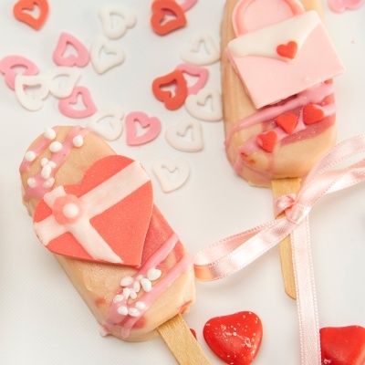 Dois gelados decorados com corações em chocolate rosa, bege e vermelho, com laço rosa e corações ao fundo.