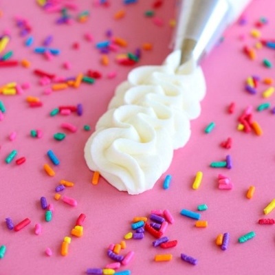 Bico de pasteleiro a aplicar creme branco sobre fundo rosa com granulados coloridos