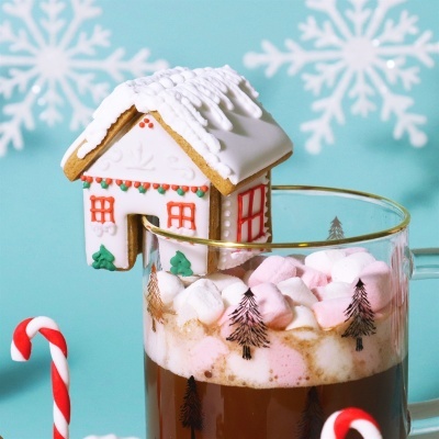 Caneca com chocolate quente e marshmallows decorada, com pequena casa de gengibre pendurada e flocos de neve no fundo
