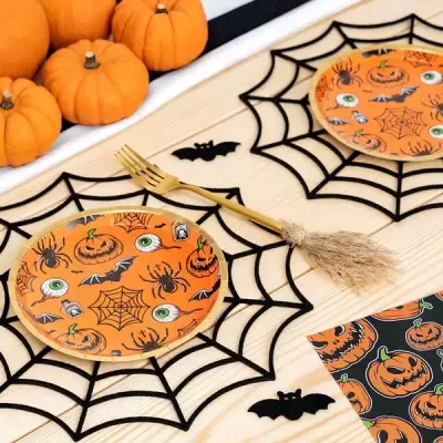 Pratos de papel decorativos de Halloween com padrões de abóboras e olhos, sobre bases de teias de aranha, com talheres dourados e mini abóboras ao lado.