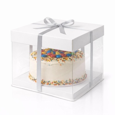 Bolo redondo branco com confeitos coloridos numa caixa transparente com tampa branca e fita cinza