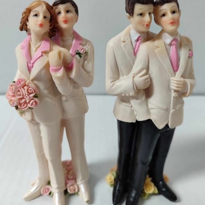 Figuras decorativas de casais do mesmo sexo, duas mulheres e dois homens, com roupas brancas e acessórios cor-de-rosa, em bases com flores.