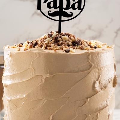 Bolo com cobertura bege, decorado com raspas de chocolate e topo com texto FELICIDADES PAPÁ em acrílico preto