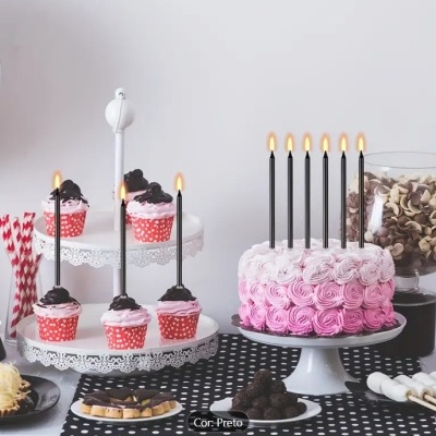 Bolo rosa com velas pretas, cupcakes com cobertura rosa e chocolate e vários doces numa mesa com toalha preta e branca.