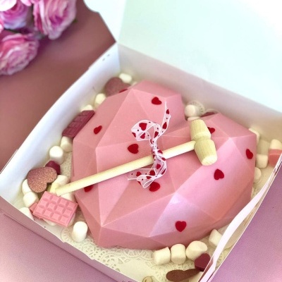 Bolo cor-de-rosa em forma de coração geométrico com malho de madeira e decoração de marshmallows e chocolates dentro de caixa branca