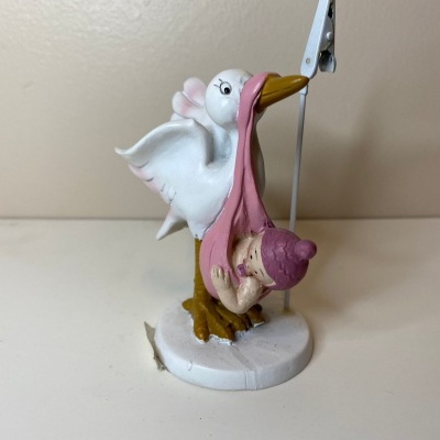 Figura decorativa de cegonha branca a transportar bebé em manta cor-de-rosa.