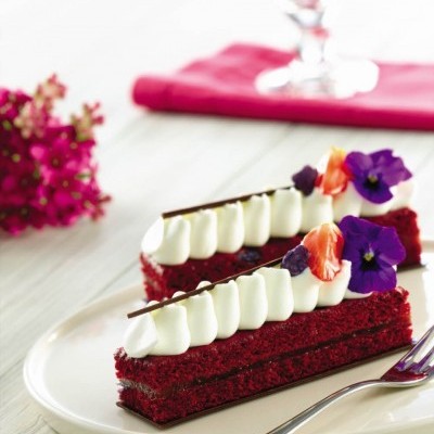 Duas fatias de bolo vermelho decoradas com cobertura branca, flores roxas e chocolate num prato branco.