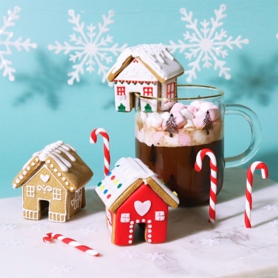 Casas de gengibre decoradas e caneca com chocolate quente e marshmallows com fundo azul e flocos de neve.