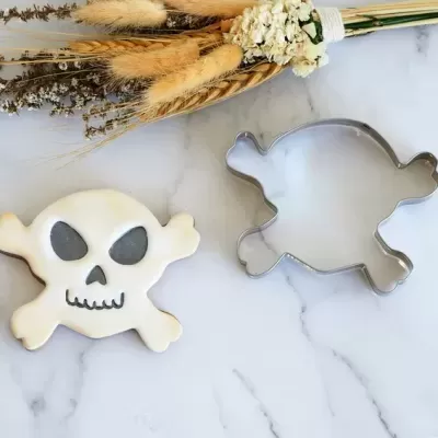 Cortador de bolachas metálico em forma de caveira com biscoito decorado à esquerda e flores secas ao fundo