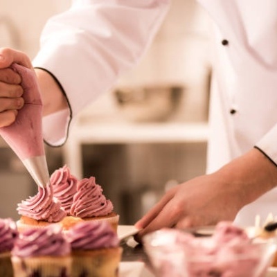 Chef a decorar bolos pequenos com cobertura rosa usando saco de pasteleiro.
