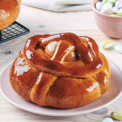 Pão doce com ovo cozido e brilho, em prato branco, com tigela de amêndoas coloridas ao fundo.