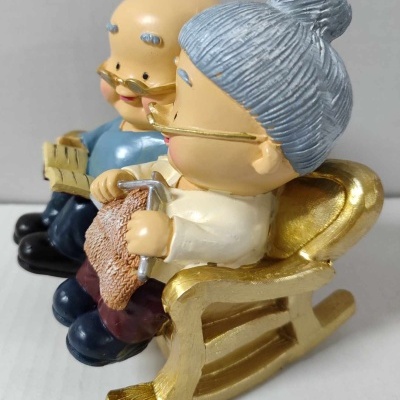 Figuras decorativas de casal de idosos a ler e tricotar numa cadeira de baloiço dourada
