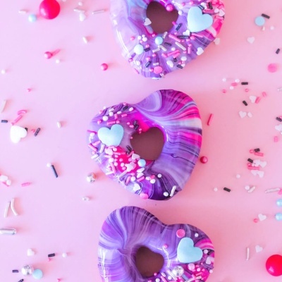 Três donuts em forma de coração decorados com glacê roxo e confeitos coloridos sobre fundo rosa