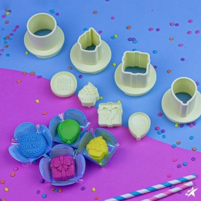 Conjunto de cortadores e carimbos para bolachas em plástico branco, coloridos, fundo azul e rosa com confetes.