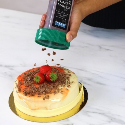 Bolo redondo com cobertura branca e laranja, morangos e raspas de chocolate sendo polvilhadas