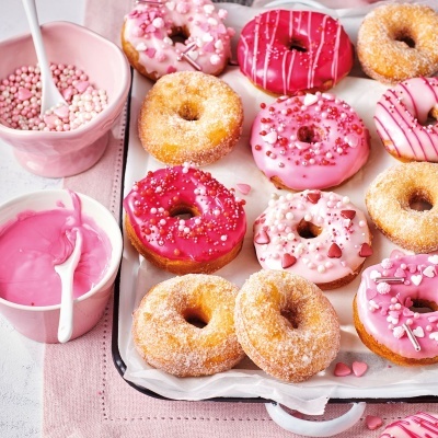 Doze donuts coloridos com coberturas decorativas em uma travessa, com taças de creme rosa ao lado