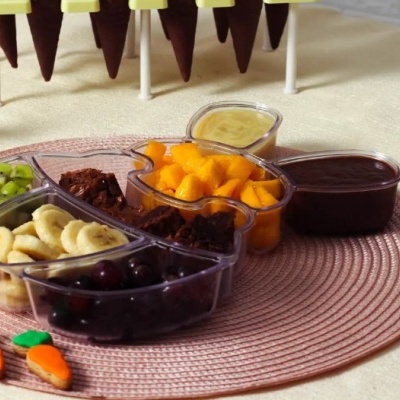 Tabuleiro de plástico transparente com frutas, brownie e molhos sobre tapete rosa