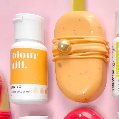 Produtos coloridos incluindo frasco com rótulo 'colour mill.' e objeto oval com pontos dourados sobre fundo rosa