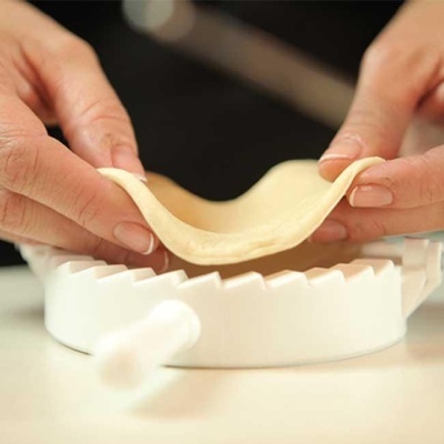 Mãos esticando massa sobre prensa branca para fazer pastéis.