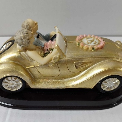 Veículo decorativo dourado com casal de bonecos sentados em base preta