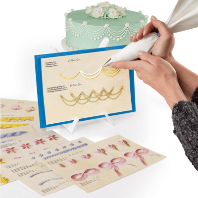 Pessoa a praticar decoração de bolos com glacê em cartão instrutivo, bolo decorado ao fundo