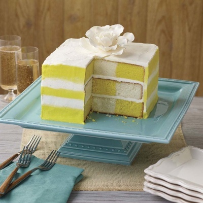 Bolo amarelo e branco decorado com flor branca, servido num prato azul com garfos e pratos ao lado.