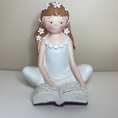 Boneca decorativa de menina sentada com vestido branco e livro aberto