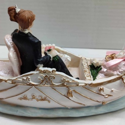 Figurina de casal num barco branco com detalhes dourados, mulher de vestido preto segurando flores