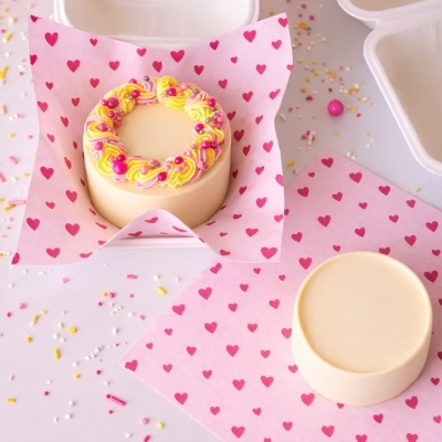 Dois bolos pequenos redondos em papel rosa com corações vermelhos, decorados com creme e confeitos coloridos.