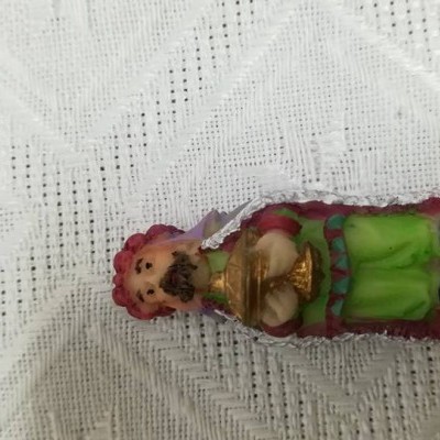 Figura decorativa pequena com roupa tradicional em verde, rosa e dourado sobre fundo branco texturizado