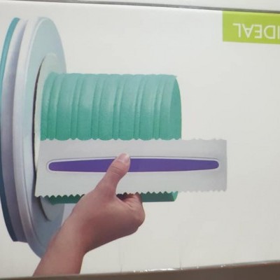 Mão segurando espátula branca com faixa roxa e anéis verdes numa superfície redonda branca