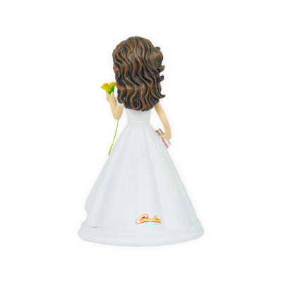 Boneca de vestido branco com cabelo castanho ondulado, segurando uma flor amarela e um livro, vista de costas.