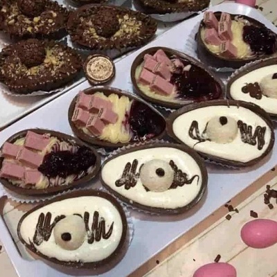 Ovos de chocolate decorados com diferentes coberturas e ingredientes variados, sobre bandejas brancas, com ovos rosa à volta.