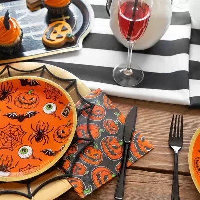 Mesa de madeira com decoração de Halloween, pratos laranja com desenhos de abóboras e aranhas, guardanapo com abóboras, talheres pretos e copo com bebida vermelha.