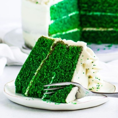 Fatia de bolo verde com creme branco em prato com garfo, bolo inteiro ao fundo