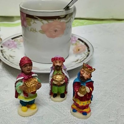 Três miniaturas coloridas dos Reis Magos em frente a chávena e pires floral sobre mesa branca.