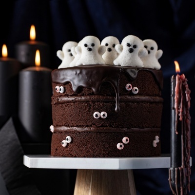 Bolo de chocolate decorado com fantasmas brancos e olhos coloridos, velas pretas acesas ao fundo.