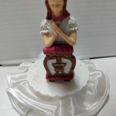 Figura decorativa de menina em oração com vestido branco e rosa ajoelhada sobre banco marrom