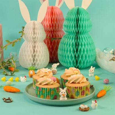 Cupcakes decorados com creme em forminhas coloridas em prato verde, rodeados por decorações de Páscoa com coelhos, cenouras e ovos sobre fundo azul claro.