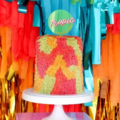 Bolo colorido com textura felpuda em suporte branco com fundo festivo de fitas coloridas