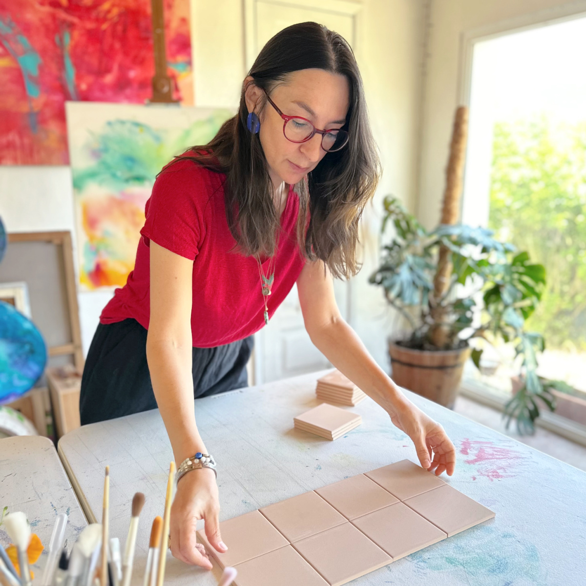 Um acaso que se tornou paixão…eu e os Azulejos! Mulher organizando azulejos quadrados de cerâmica bege sobre mesa com pincéis e quadros coloridos ao fundo
