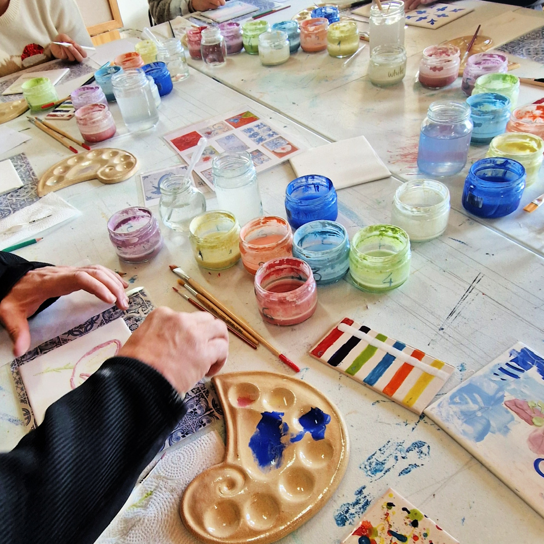 Tile Painting Workshop Mesa de pintura com frascos de tinta, pincéis e azulejos decorativos.