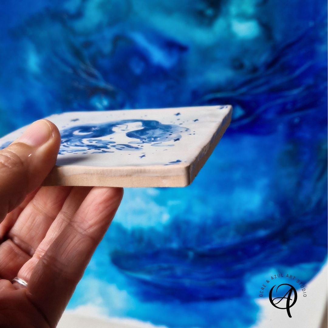 Azulejo Coleção Deusas e Luas Azulejo branco com padrão azul pintado à mão segurado por uma mão, fundo azul abstrato, logótipo do estúdio de arte