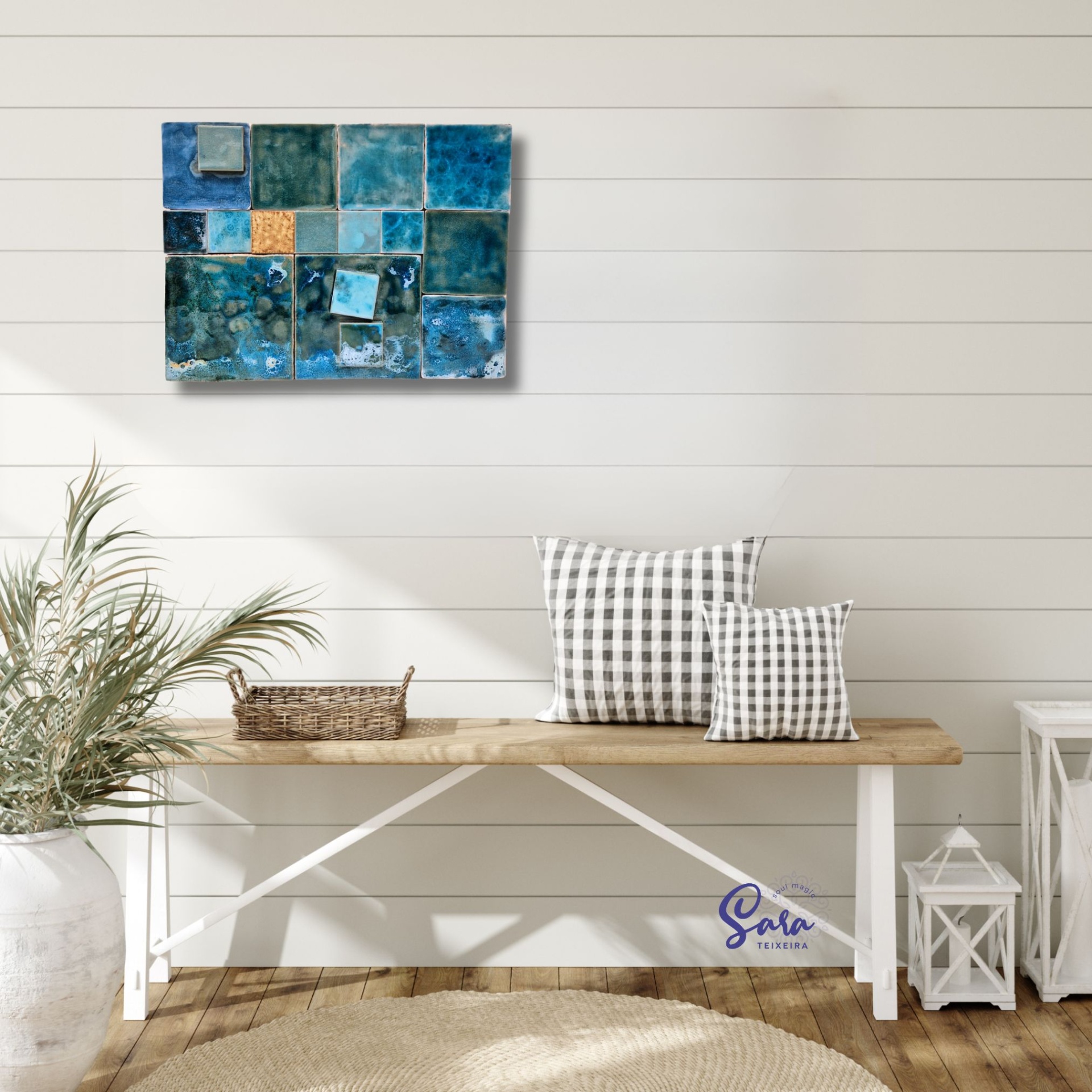 Painel de Azulejo Sinfonia em Turquesa Conjunto de azulejos azuis pendurado numa parede branca com mesa e almofadas quadriculadas.