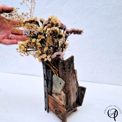 Vaso de madeira reciclada com arranjo de flores secas e mão com anel ao lado
