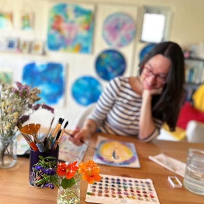 Mesa com pincéis, paleta de aquarela, flores num jarro e mulher a pintar com quadros de arte colorida na parede