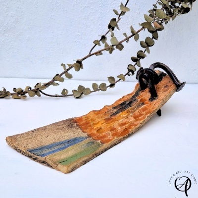Escultura cerâmica de prancha de surf com peças metálicas pretas e ramo seco ao fundo.