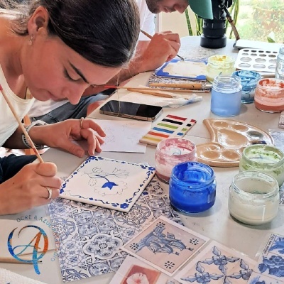 Pessoa a pintar azulejo branco com padrão floral azul e materiais de pintura à volta