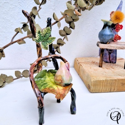 Mini cadeira decorativa de cerâmica com jarra, folhas pintadas, sobre mesa branca com galhos secos e suporte de madeira